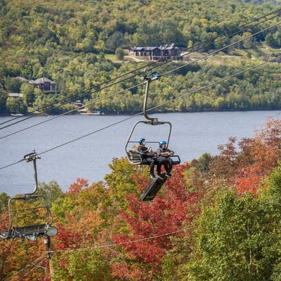 A panoramic of customers riding the Luge chairlift at Mont Tremblant with the lake behind.