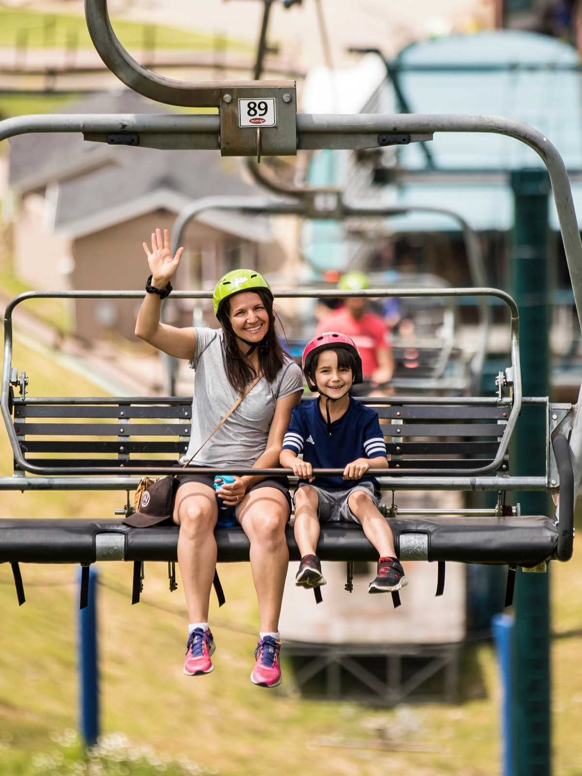 A mother and son smile and wave as they ride the chairlift to the top of the tracks.