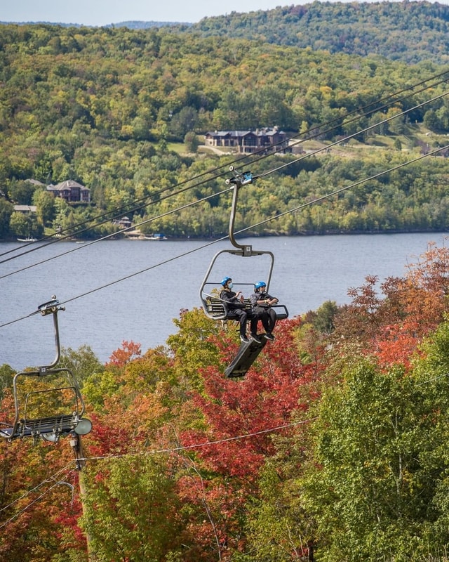 A panoramic of customers riding the Luge chairlift at Mont Tremblant with the lake behind.